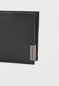 Zwarte leren portefeuille met een gestructureerde textuur, voorzien van een klein zilveren metalen label met de gravure "Tommy Hilfiger" in de hoek, tegen een effen achtergrond.