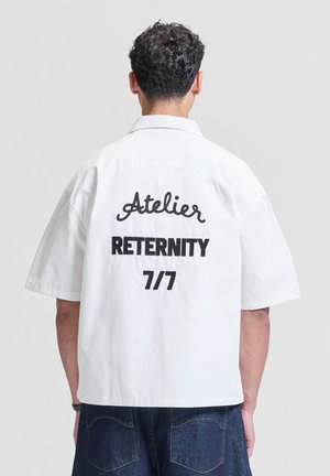 Mężczyzna z kręconymi włosami, ubrany w białą koszulę z krótkim rękawem, na której z tyłu nadrukowano "Atelier RETERNITY 7/7", oraz ciemnoniebieskie dżinsy.