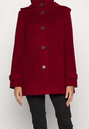 Vrouw draagt een knielange rode wollen jas met zwarte knopen, manchetten met riempjes en een hoge kraag, gecombineerd met een zwarte broek.