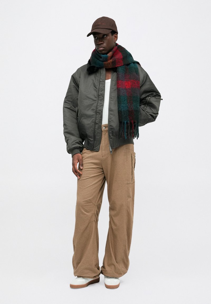 Veste bomber grise, pantalon beige à jambe large, chemise blanche, écharpe à carreaux rouges et verts, casquette marron, baskets blanches avec semelles beiges.
