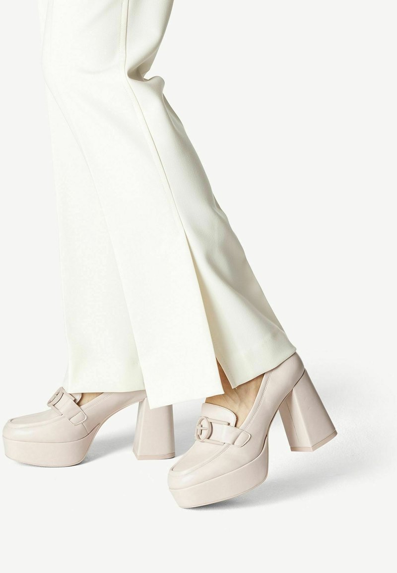 Femme portant un pantalon blanc à jambes larges avec des fentes latérales et des mocassins beiges à plateforme et talons hauts sur fond blanc.