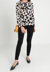 Blouse à manches longues avec un motif abstrait noir et blanc, associée à un pantalon noir ajusté et des ballerines noires pointues. Accessoirisée d'un sac jaune.