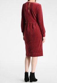 Bordowa sukienka z miękkiego, teksturowanego materiału, z długimi rękawami, dopasowaną talią oraz wycięciem w kształcie łezki z tyłu, sięgająca do połowy łydki.