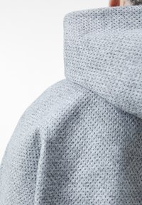 Lys grå hettegenser laget av teksturert stoff med et maske-mønster. Den har høy krage og en strukturert, komfortabel design.