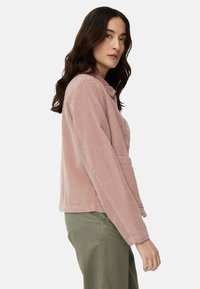 Różowa kurtka sztruksowa z kołnierzem, zapięciem na guziki i przednią kieszenią; charakteryzuje się fakturowaną tkaniną i luźnym krojem. W zestawie z zielonymi spodniami.