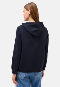 Kvinde med langt brunt hår iført en marineblå hættetrøje og lyseblå jeans, stående med ryggen til mod en ensfarvet lys baggrund.