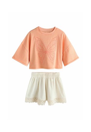 Gyerek, aki egy őszibarackszínű, túlméretezett inget visel pillangó hímzéssel és fehér texturált rövidnadrágot csipkés szegéllyel, sima háttér előtt áll.