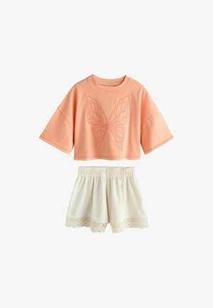 Niño con camisa extragrande color melocotón con bordado de mariposa y pantalones cortos blancos texturizados con ribete de encaje, de pie frente a un fondo liso.