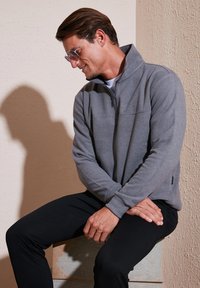 Pullover grigio in pile con colletto alto, zip e colore uniforme. Il modello indossa pantaloni neri e posa seduto su uno sfondo beige texturizzato.