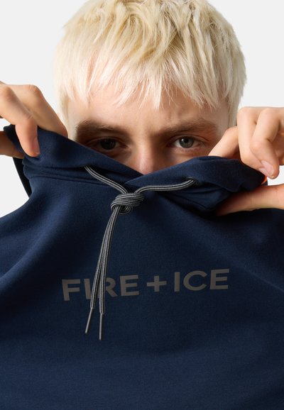 Mladý člověk s krátkými blond vlasy si zvedá námořnicky modrou kapuci s nápisem "FIRE + ICE" šedým písmem a viditelným stahovacím šňůrkem.