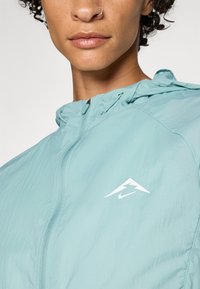 Veste légère à capuche en teal pastel, dotée d'une fermeture éclair et d'un logo blanc sur la poitrine. Texture ripstop avec une finition lisse.
