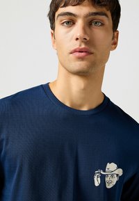 Camiseta de manga corta azul marino hecha de tejido suave. Presenta un gráfico blanco de un vaquero con sombrero e iniciales en el lado izquierdo.