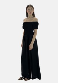 Sort off-shoulder kjole med smock-detalje på overdelen og lang, fløjlsagtig nederdel. Glat tekstur, ankellang, parret med sorte sandaler.