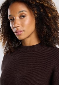 Brown ribbed knit sweater with a high neckline, featuring textured fabric. Model has curly hair and natural makeup, focusing on the garment.
