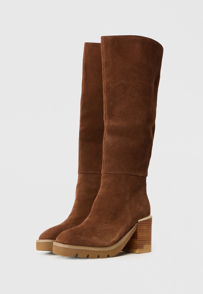 Botas de ante marrón hasta la rodilla con un tacón de madera grueso, punta redondeada y acabado texturizado. Costuras notables a lo largo del eje de la bota.