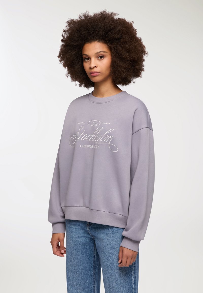 Jasnofioletowy sweter z luźnym krojem, z ściągaczami na rękawach i u dołu. Z przodu znajduje się dekoracyjny biały napis "Stockholm Leisure Club."
