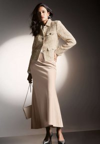 Beige tweed jack met twee voorzakken en knopen, gecombineerd met een lange, gladde beige rok en zwarte hoge hakken.