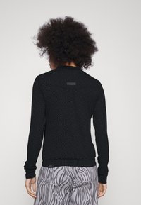 Svart långärmad topp med ett subtilt prickmönster, med en rund halsringning och ribbade ärmslut. Baksidan har en liten detaljerad varumärkesetikett.