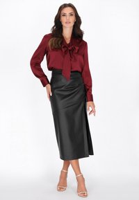 Burgundowa satynowa bluzka z kołnierzykiem w kształcie kokardy, zestawiona z czarną, ekologiczną skórzaną spódnicą w stylu owijanym z bocznym rozcięciem i gładką fakturą. Przezroczyste sandały.