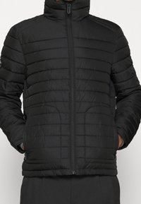 Chaqueta acolchada negra con collar alto, diseño de acolchado horizontal y cierre de cremallera. Cuenta con bolsillos laterales y puños elásticos para un ajuste ceñido.