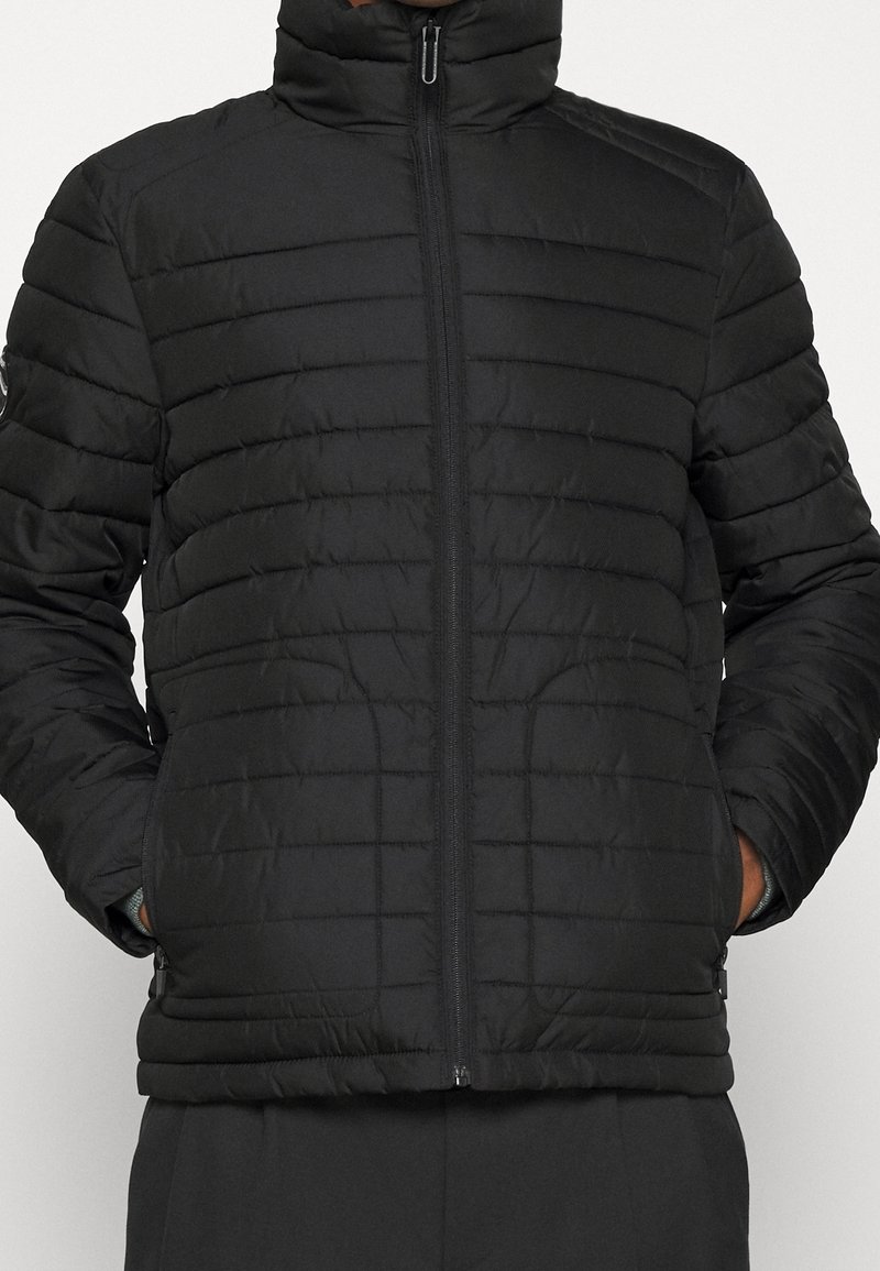 Chaqueta acolchada negra con collar alto, diseño de acolchado horizontal y cierre de cremallera. Cuenta con bolsillos laterales y puños elásticos para un ajuste ceñido.