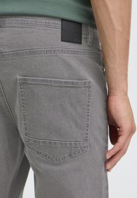 Szürke farmernadrág hátsó zsebbel és fekete bőr címkével. Az anyag puhanak tűnik, finom textúrával.