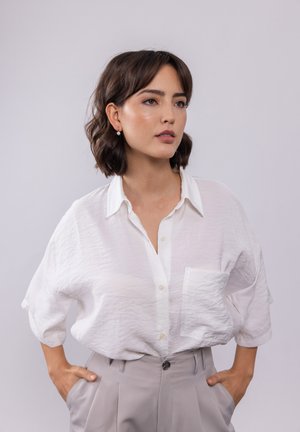 Witte, gestructureerde button-up blouse met een zak, gecombineerd met lichtgrijze high-waisted broek. Het model heeft kort, los, golvend haar.