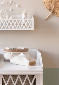 Trocador branco com uma prateleira em padrão de diamante, sacos de armazenamento às riscas bege e um leque decorativo de parede feito de materiais naturais.