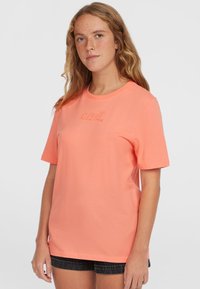 Camiseta de algodón color coral con mangas cortas, cuello redondo y logo bordado en el pecho. La tela tiene una textura suave.