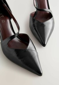 Pantofole negre din piele cu un design cu vârf ascuțit, având bretele încrucișate peste o parte deschisă și o textură netedă, lucioasă.