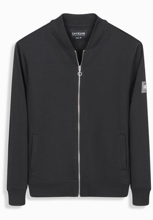 Chaqueta negra de cremallera con tejido suave, con cuello y puños acanalados. Cuenta con bolsillos laterales y un parche con el logo en la manga. Talla mediana.