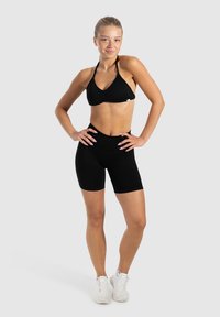 Zwarte sport-bh met halternek ontwerp en plooidetail, gecombineerd met zwarte shorts met hoge taille. Model draagt witte sportschoenen.