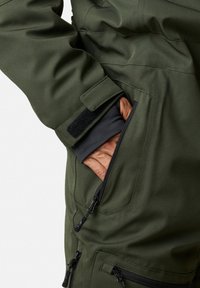 Oliwkowa kurtka z teksturowanej tkaniny, z zapinaną na zamek kieszenią i zabezpieczonym zapięciem na mankiet. Wyraźna kontrastowa czarna podszewka.