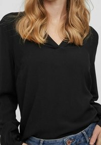 Blusa nera a maniche lunghe con scollatura a V. Tessuto liscio con leggero lucido. Abbinata a jeans blu chiaro, con un dettaglio di bottoni.