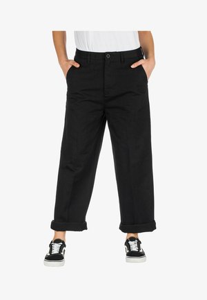 Melni, plata kāju bikses ar gludu tekstūru, ar pogu aizdari, sānu kabatām un sarullētajiem piedurkņu galiem, apvienotas ar melnbaltām kedām.