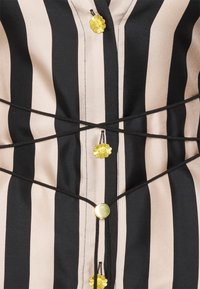 Hemd mit Knöpfen in schwarz-beigen Streifen. Verfügt über gelbe Blumenverzierungen und ein schräges schwarzes Zugband-Detail. Glatte Textur.