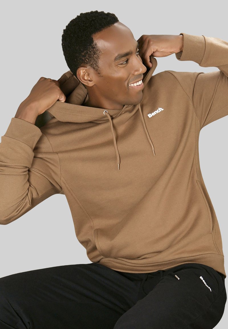 Brauner Kapuzenpullover mit Kordelzug, ausgestattet mit einer Vordertasche und dem weißen "Bench."-Logo. Tragen mit schwarzen Hosen vor einem grauen Hintergrund.