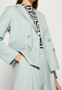 Vaaleansininen räätälöity bleiseri, jossa on kaksoisnapitus, lovellut kaulukset ja teksturoitu kangas, yhdistetty mustavalkoiseen seepra-kuvioiseen toppiin alla.