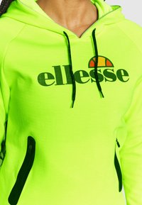 Sudadera verde neón con un gran logo negro de "ellesse" y un acento de color en la parte superior. Incluye bolsillos con cremallera negra y cordones de contraste.