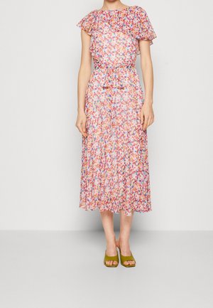 Kobieta w wielokolorowej sukience midi w kwiaty z bufiastymi rękawami i wiązaniem w talii, w połączeniu z oliwkowo zielonymi sandałami na obcasie, stojąca na jednolitym tle.