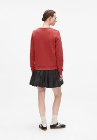 Felpa rossa di cotone con maniche lunghe, polsini e orlo a coste, abbinata a una gonna nera plissettata. Sul retro presenta un logo stampato vicino al collo.