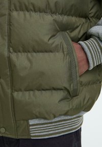 Maslinasto zeleni pufasti kaput s horizontalno šivenim dijelovima, rebrastim sivim i tamnozelenim manžetama te bočnom džepom. Glatka sjajna tekstura.