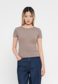 Femme jeune avec des cheveux blonds ombrés lisses, portant un haut à manches courtes en taupe côtelé et un jean taille haute bleu, se tenant avec une main dans la poche.