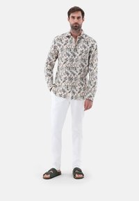 Dalla camicia a maniche lunghe con uno sfondo chiaro presenta un motivo di foglie multicolori, abbinata a dei pantaloni bianchi e sandali scuri.