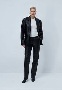 Sieviete ar blondiem matiem, ģērbta melnā ādas žaketē, melnās ādas biksēs, baltā pieguļošā topā, ar apaļām auskaru gredzeniem un melnām augstpapēžu kurpēm ar norādītu purngalu.