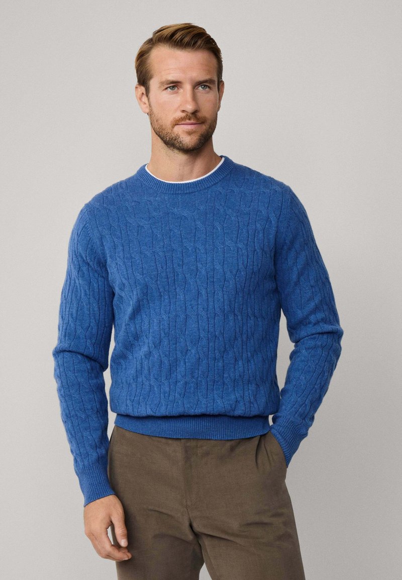 Suéter azul de punto trenzado con cuello redondo, patrón texturizado, puños y dobladillo acanalados. Combinado con pantalones marrones, creando un look en capas.