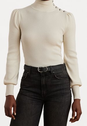 Pull à col roulé côtelé crème avec épaules bouffantes et boutons décoratifs sur un côté, associé à un jean noir taille haute et une ceinture.