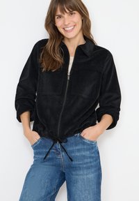 Czarna kurtka sztruksowa zapinana na zamek, z dwoma kieszeniami na piersiach, podwiniętymi rękawami oraz regulowanym sznurkiem w talii, w połączeniu z niebieskimi dżinsami.