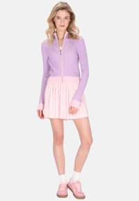 orgonalila, bordázott cipzáras pulóver rózsaszín derékpánttal, világos rózsaszín pliszírozott szoknyával párosítva. Rózsaszín tornacipővel és fehér zoknival viselve.