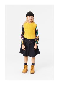 Rumena pletena brezrokavna majica, cvetlična srajca z dolgimi rokavi, temno denim krilo s gumbi, svetlo rjave gležnjarje in črna pletena kapa.